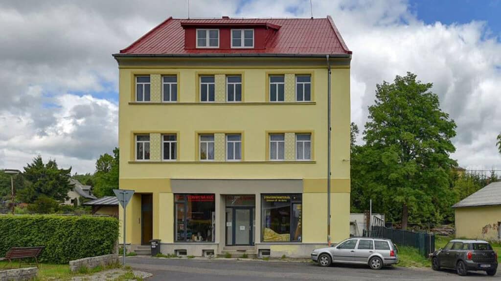 Soubor s penzionem v centru města, třípatrová budova s žlutým fasádou, komerční provoz v přízemí, parkování před objektem, okolí s stromy a sousedními domy, ideální pro ubytování a relaxaci.